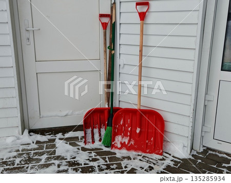 Red brush for sweeping snow from the floor. Shovels for snow removal near the utility room Red brush for sweeping snow from the floor. Shovels for snow removal near the utility room 135285934