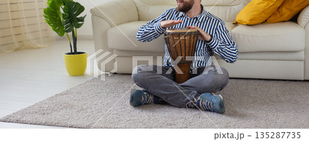 Man playing hand drum while sitting on sofa in living room. Banner with copy space. Rhythm practice, percussion hobby, and relaxed music session at home. Man playing hand drum while sitting on sofa in living room. Banner with copy space. Rhythm practice, percussion hobby, and relaxed music session at home. 135287735
