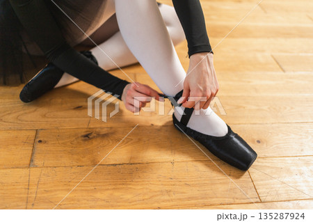 Ballerina puts on pointe shoes on leg in dance class. Classical ballet dancer woman in dance studio. Ballerina putting on tying classical pointe shoes for dance training. Ballet school concept Ballerina puts on pointe shoes on leg in dance class. Classical ballet dancer woman in dance studio. Ballerina putting on tying classical pointe shoes for dance training. Ballet school concept 135287924