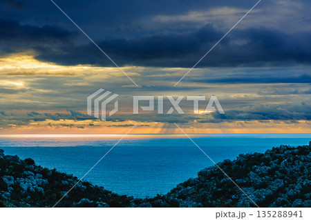 Serene Sunset Over Calm Sea With Rocky Hills Framing The Distant Golden Horizon 135288941