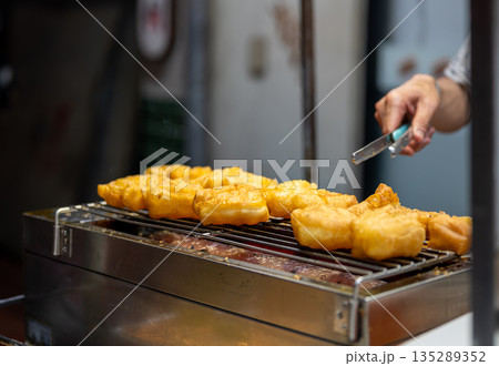 Close-up view of a street food cook turning freshly fried patongo dough with metal tongs, capturing the hot oil, golden texture, and authentic Thai street cuisine atmosphere. Close-up view of a street food cook turning freshly fried patongo dough with metal tongs, capturing the hot oil, golden texture, and authentic Thai street cuisine atmosphere. 135289352