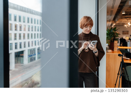 Focused female entrepreneur standing near large window in modern office, holding smartphone and reading information. Calm expression, business work environment, architectural background. Focused female entrepreneur standing near large window in modern office, holding smartphone and reading information. Calm expression, business work environment, architectural background. 135290303