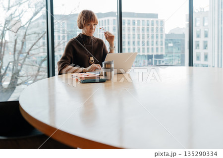 Stylish business woman with notebook, laptop and coffee cup working alone at big around table on laptop while sitting in modern office near window Stylish business woman with notebook, laptop and coffee cup working alone at big around table on laptop while sitting in modern office near window 135290334