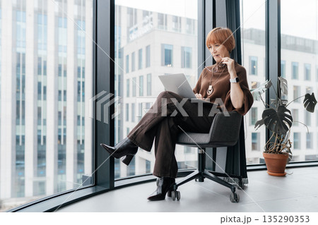 White woman aged 49 sitting on office chair near large window, full body shot, laptop on knees. Independent researcher or advisor reviewing, modern business building outside, reset during workday White woman aged 49 sitting on office chair near large window, full body shot, laptop on knees. Independent researcher or advisor reviewing, modern business building outside, reset during workday 135290353