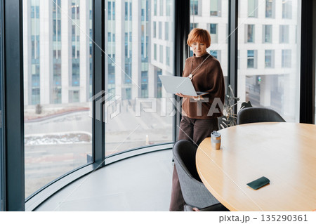 Full body view of independent female near meeting table and panoramic window, with laptop during work break. Modern office space, urban outside, quiet focus, professional routine, copy space 135290361