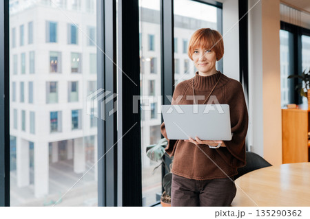 Portrait of confident female consultant 50s with short red hair holding laptop near office window. Calm smile, digital expertise, independent work style, modern coworking interior, copy space 135290362