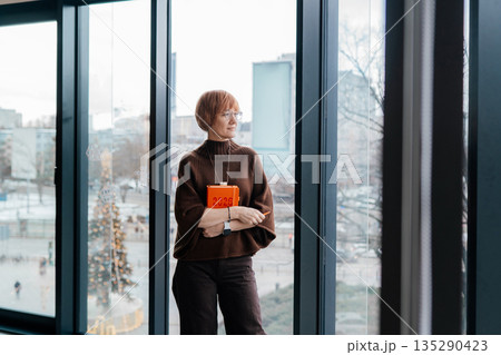 Middle aged confident woman standing by panoramic window in modern office. Concept of female leadership, independent work, decision, business planning, financial, experience, professional lifestyle Middle aged confident woman standing by panoramic window in modern office. Concept of female leadership, independent work, decision, business planning, financial, experience, professional lifestyle 135290423