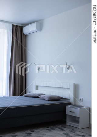 Light bedroom with bed near window and air conditioner above the bed. Bedroom interior. Light bedroom with bed near window and air conditioner above the bed. Bedroom interior. 135290701