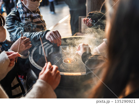 Close focus on tourists' hands burning the small incenses with smoke inside the big pot. 135290921
