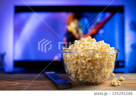 A glass bowl of popcorn and remote control in the background the TV works. Evening cozy watching a movie or TV series at home A glass bowl of popcorn and remote control in the background the TV works. Evening cozy watching a movie or TV series at home 135291288