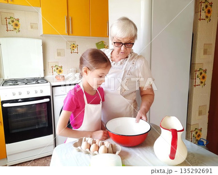 A grandmother and granddaughter stand at a table with a bowl, eggs, and a pitcher. A grandmother and granddaughter stand at a table with a bowl, eggs, and a pitcher. 135292691