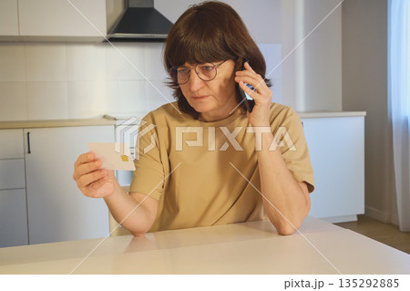 Woman holding a credit card and talking on a mobile phone in a kitchen at home Woman holding a credit card and talking on a mobile phone in a kitchen at home 135292885