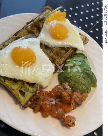 Breakfast plate with eggs on waffles, avocado slices, and tomato salad on white polka dot table 135293756