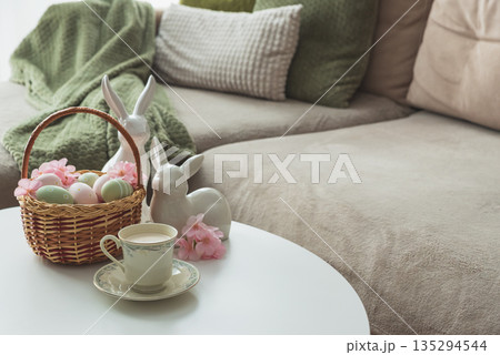 Easter Basket with Ester Eggs, Bunny Figurines and coffee cup on White Table intoCozy Living Room 135294544