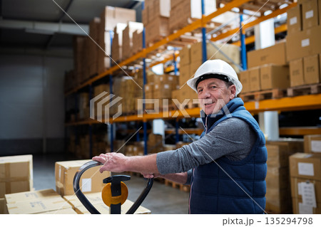 Older male warehouse worker pulling a pallet truck. Older male warehouse worker pulling a pallet truck. 135294798
