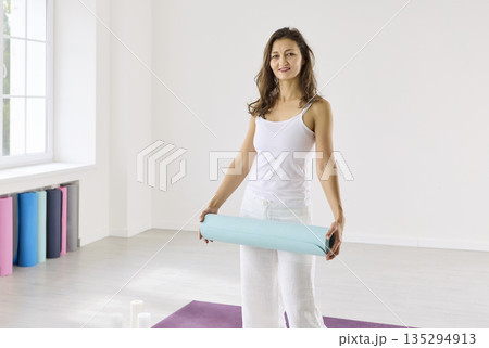 Woman in white holding yoga mat on purple mat in bright studio with soft light Woman in white holding yoga mat on purple mat in bright studio with soft light 135294913