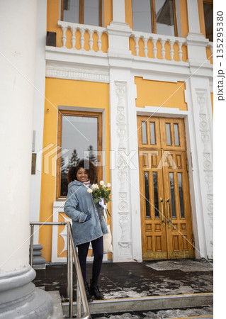 African american Woman holding bouquet of white roses in winter sunlight outdoors. Joy, tenderness, and individuality expressed through urban winter lifestyle and seasonal atmosphere. African american Woman holding bouquet of white roses in winter sunlight outdoors. Joy, tenderness, and individuality expressed through urban winter lifestyle and seasonal atmosphere. 135295380