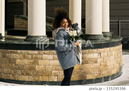 African american Woman holding bouquet of white roses in winter sunlight outdoors. Joy, tenderness, and individuality expressed through urban winter lifestyle and seasonal atmosphere. African american Woman holding bouquet of white roses in winter sunlight outdoors. Joy, tenderness, and individuality expressed through urban winter lifestyle and seasonal atmosphere. 135295386
