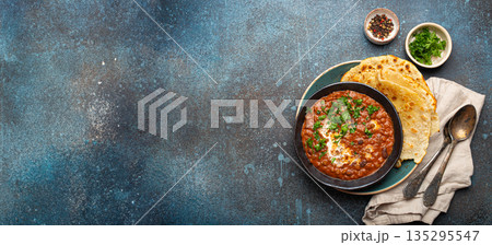 Traditional Indian Punjabi dish Dal makhani with lentils and beans in black bowl served with naan flat bread, fresh cilantro and two spoons on blue concrete rustic table top view. Space for text Traditional Indian Punjabi dish Dal makhani with lentils and beans in black bowl served with naan flat bread, fresh cilantro and two spoons on blue concrete rustic table top view. Space for text 135295547