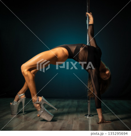 Pole dancer arching back on pole under dramatic studio lighting, high heels and sheer costume highlighting toned muscles and graceful curve, expressive silhouette conveying strength and sensual 135295694