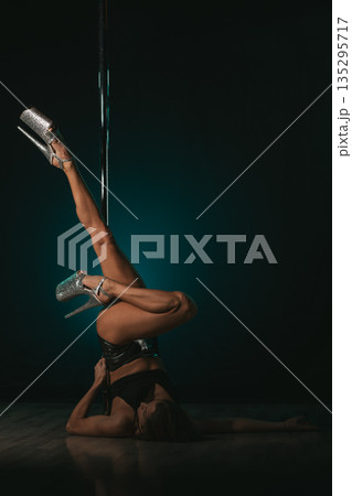 Floor inversion with legs extended upward gripping pole, heels sparkling against moody backdrop, core strength and focused expression conveying disciplined training and pole dance performance energy 135295717
