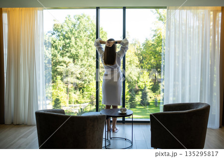 Slim woman in white bath robe standing at the window in a hotel suit 135295817