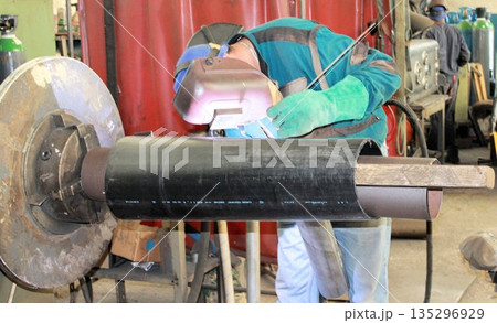 Industrial worker with protective helmet and green gloves performing TIG welding on a large black metal pipe. Skilled labor in a mechanical engineering factory. Industrial worker with protective helmet and green gloves performing TIG welding on a large black metal pipe. Skilled labor in a mechanical engineering factory. 135296929