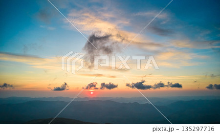 Sun sets behind rolling hills, casting warm, golden glow across sky. Wispy clouds, tinged with hues of orange and yellow, stretch across deepening blue sky. Serene and expansive landscape. Sun sets behind rolling hills, casting warm, golden glow across sky. Wispy clouds, tinged with hues of orange and yellow, stretch across deepening blue sky. Serene and expansive landscape. 135297716