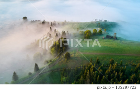 Aerial view of foggy morning in countryside. Mist envelopes lush green hills, dotted with trees and few houses, creating serene and mystical atmosphere as sunlight filters through. Aerial view of foggy morning in countryside. Mist envelopes lush green hills, dotted with trees and few houses, creating serene and mystical atmosphere as sunlight filters through. 135297722