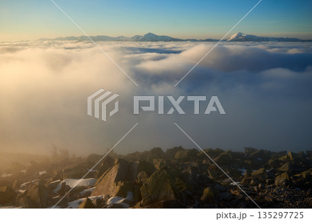 Sunrise over snow-covered mountain peak. Warm golden light of rising sun bathes clouds in soft hues of orange and pink. Snow and scattered rocks in foreground add texture and depth to scene. Sunrise over snow-covered mountain peak. Warm golden light of rising sun bathes clouds in soft hues of orange and pink. Snow and scattered rocks in foreground add texture and depth to scene. 135297725