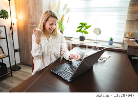 Young woman celebrates success while working on laptop at home office Young woman celebrates success while working on laptop at home office 135297947