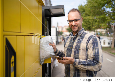 Man using a package delivery locker on a sunny day in an urban setting while checking his phone 135298034
