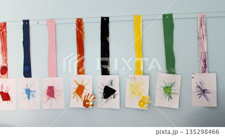 A row of creative handmade artworks by preschool children displayed on a light blue classroom wall. The crafts include colorful painted strips and finger-painted handprints on white paper, showcasing A row of creative handmade artworks by preschool children displayed on a light blue classroom wall. The crafts include colorful painted strips and finger-painted handprints on white paper, showcasing 135298466