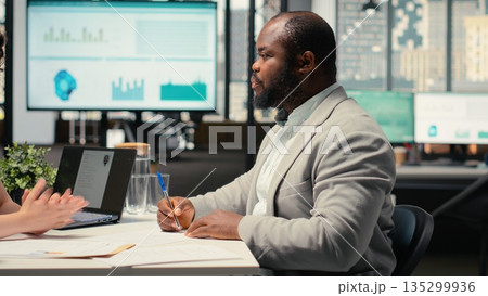 African american new hire signing work contract terms to start new job, receiving approval for onboarding within enterprise. Recruiter finalizing hiring process with legal paperwork. 135299936
