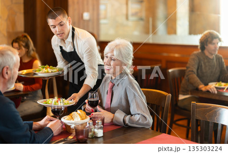 In cafe, guy waiter serves catering establishments mature couple visitor, puts food on table. 135303224