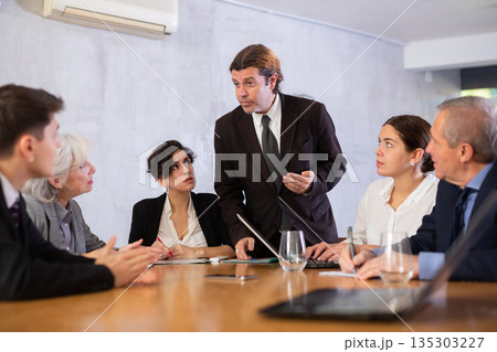 Displeased male manager scolding at young, adult, mature office workers sitting with reports and laptops at table in meeting room Displeased male manager scolding at young, adult, mature office workers sitting with reports and laptops at table in meeting room 135303227