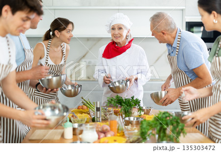 Smiling old woman chef of cooking course teaching attendees how to mix sauce 135303472