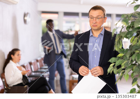 Upset middle-aged man standing in waiting room with his back to person who is shouting at him Upset middle-aged man standing in waiting room with his back to person who is shouting at him 135303487