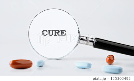 The word Cure is magnified by a magnifying glass resting near pills of different sizes and colors scattered on a clean, white backdrop in a studio setting 135303493