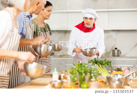 Young guy cook teaches to cook group of people Young guy cook teaches to cook group of people 135303537