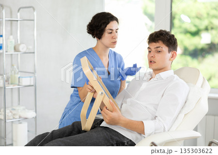 Man sits in chair at a doctor appointment with beautician. Woman doing facial examination before surgery 135303622