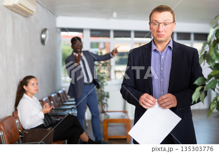 Upset middle-aged man standing in waiting room with his back to person who is shouting at him Upset middle-aged man standing in waiting room with his back to person who is shouting at him 135303776