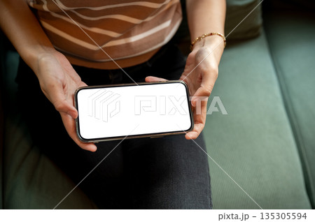 Close up of woman's hands holding blank screen phone playing or watching while sitting on green sofa Close up of woman's hands holding blank screen phone playing or watching while sitting on green sofa 135305594