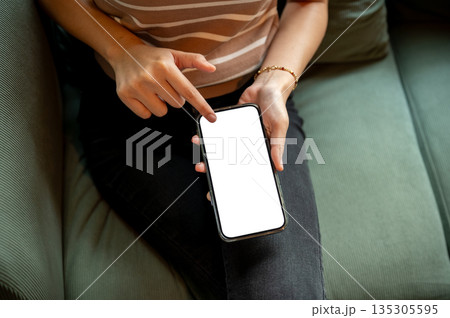Close up of woman hand holding and touching on blank screen smartphone while sitting on green sofa. Close up of woman hand holding and touching on blank screen smartphone while sitting on green sofa. 135305595