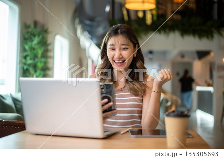 Asian woman holding phone raising a fist in happy or excited over laptop on table in cafe or canteen Asian woman holding phone raising a fist in happy or excited over laptop on table in cafe or canteen 135305693