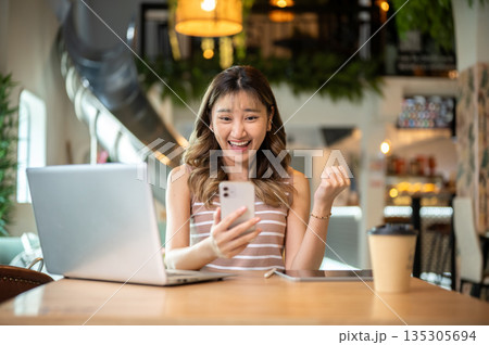 Asian woman holding phone raising fist in happy or excited aside laptop on table in cafe or canteen. 135305694