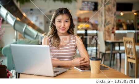Pretty asian woman sitting with tablet and coffee beside laptop on wooden table in a cafe or canteen Pretty asian woman sitting with tablet and coffee beside laptop on wooden table in a cafe or canteen 135305701
