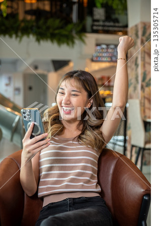 Asian woman raising a hand as looking at smartphone while sitting on brown sofa in cafe or canteen. Asian woman raising a hand as looking at smartphone while sitting on brown sofa in cafe or canteen. 135305714