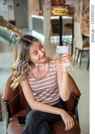 Smiling asian woman holding and looking at credit card while sits on brown sofa in cafe or canteen. Smiling asian woman holding and looking at credit card while sits on brown sofa in cafe or canteen. 135305722