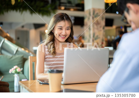 Asian woman using and looking at laptop while sitting with a man at wooden table in cafe or canteen. 135305780